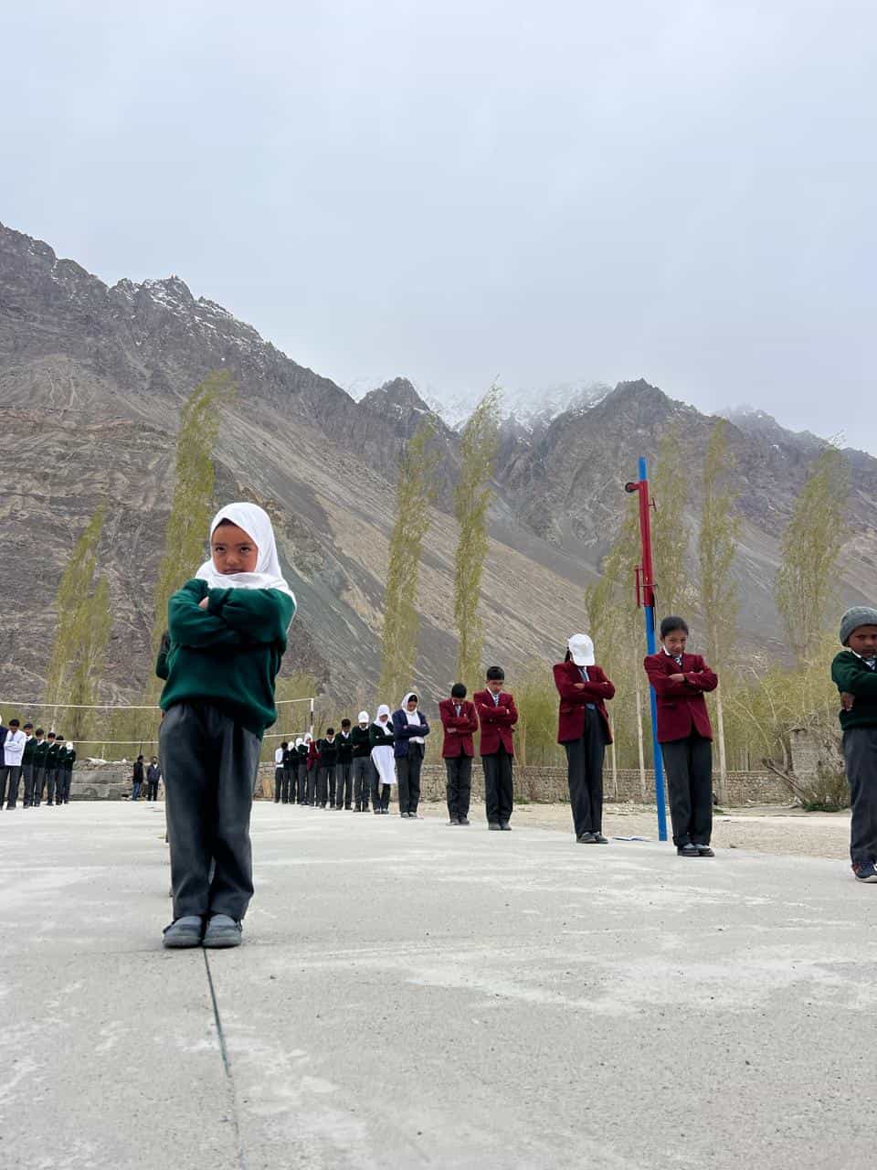 Students standing in spaced rows during morning assembly with the Nubra mountains in the background